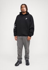 Felpa nera con cappuccio e tasca frontale, caratterizzata da un logo di ragno bianco e un design di ragnatela sulla manica sinistra. Indossata con pantaloni cargo grigi e sneaker nere.