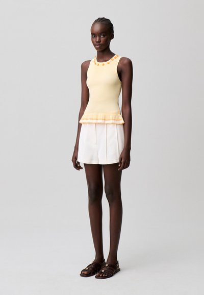 Mujer con una camiseta amarilla sin mangas con escote floral y dobladillo con volantes, pantalones cortos blancos y sandalias marrón oscuro, de pie frente a un fondo gris.