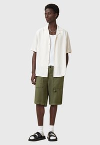 Camicia bianca a maniche corte con motivo a scacchi chiari; canotta bianca; pantaloni cargo verdi con tasche; sandali neri con dettagli lucidi; calzini alla caviglia.
