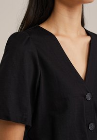 Črna kratkohlavna obleka z V-izrezom, ki ima teksturirano tkanino in tri gumbe spredaj.