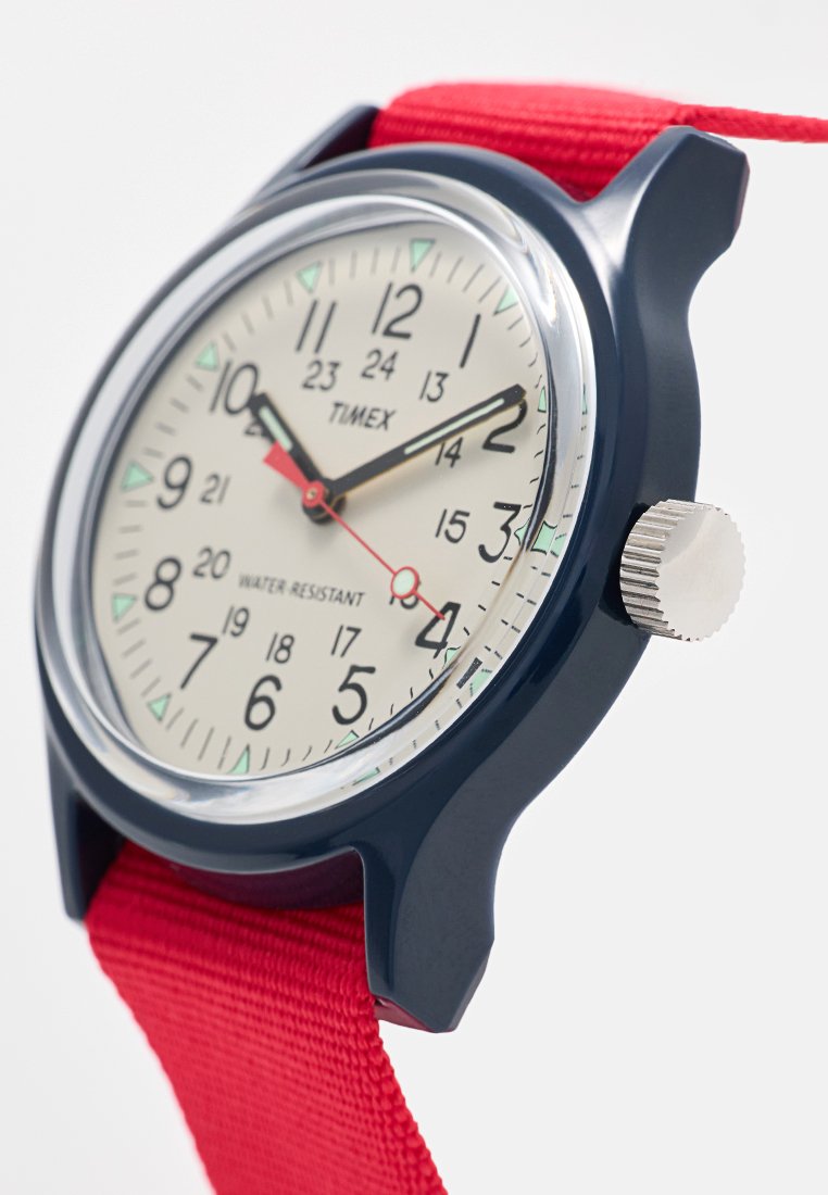 Reloj analógico Timex redondo con esfera blanca, manecillas negras y rojas, números negros, caja azul marino, corona plateada y correa de tela roja.