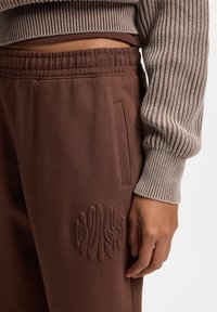 Brune joggingbukser lavet af blødt stof med en tekstureret finish, med et broderet design på låret og sidelommen.