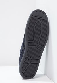 Sula av en mörk sneaker med mönstrad sula och G-STAR RAW-logotyp, vinklad mot en vit vägg och golvbakgrund.