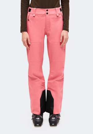 Personne portant un pantalon de ski rose vif, des guêtres noires aux chevilles et des chaussures de ski, debout devant un fond uni.