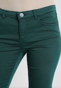 Los jeans de mezclilla verdes cuentan con una textura suave, un ajuste slim, un diseño de cinco bolsillos y un cierre frontal con botón. También se puede ver la costura a lo largo de las costuras.