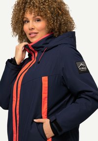 Marinblå jacka med rosa detaljer, dragkedja fram, justerbar huva och logotypplåster. Har en figurnära design och framfickor.