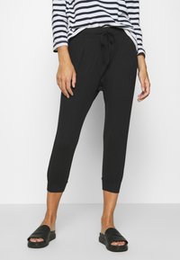 Personne portant un pantalon de jogging noir court avec une taille à cordon et des sandales noires à enfiler, associé à un haut à manches longues rayé blanc et marine.
