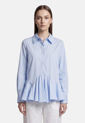 Donna con capelli castani lisci indossa una camicetta azzurro chiaro con bottoni e orlo svasato e arricciato e pantaloni bianchi, in piedi su sfondo bianco.