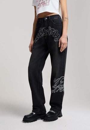 Person trägt schwarze Jeans mit weißem gotischem Schriftzug "Maldrady" und Drachengrafiken, kombiniert mit schwarzen klobigen Schuhen und einem kurzen weißen Oberteil.