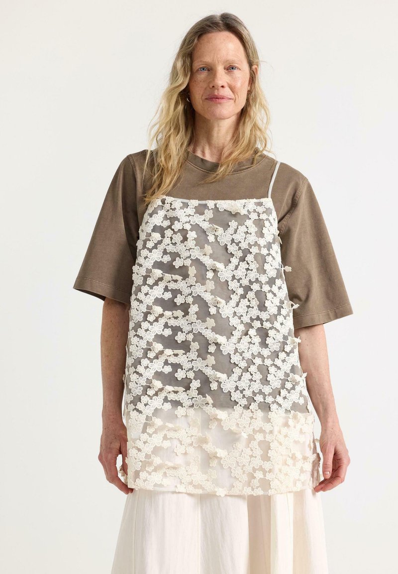 Kvinde med langt blondt hår iført brun oversized kortærmet skjorte lagdelt med gennemsigtig hvid blomsterblondekjole og hvid nederdel mod hvid baggrund.