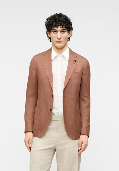 Blazer en lin marron avec deux boutons et une poche, associé à une chemise blanche et un pantalon beige clair, présentant un design minimaliste.