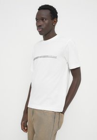 Νεαρός άνδρας που φοράει άσπρο κοντομάνικο μπλουζάκι με μαύρες ρίγες και κείμενο, μπεζ παντελόνι, στέκεται μπροστά σε ανοιχτόχρωμο απλό φόντο.