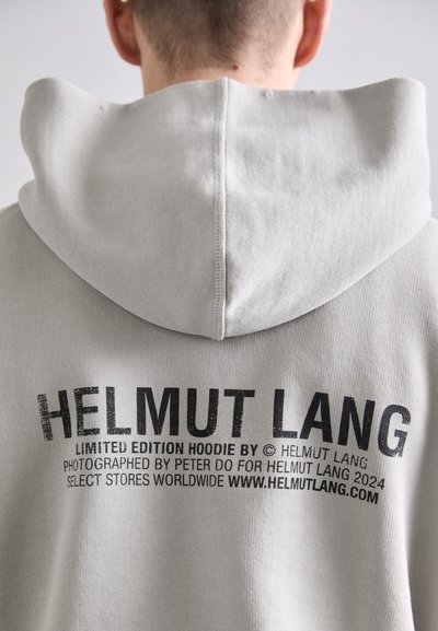 Jasnoszara bluza z kapturem z tyłu z grafiką, która głosi "HELMUT LANG" w pogrubionym czarnym napisie. Miękka tkanina z kapturem i prążkowanymi detalami.