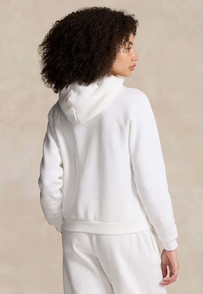 Femme aux cheveux bouclés portant un sweat à capuche blanc et un pantalon blanc, vue de dos sur un fond beige uni.