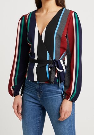 Donna che indossa una camicetta avvolgente a righe verticali multicolori con maniche lunghe e jeans blu, in piedi con le mani rilassate lungo i fianchi.