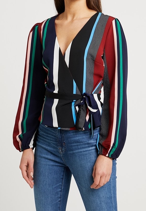 Femme portant un chemisier cache-cœur rayé vertical multicolore à manches longues et un jean bleu, debout avec les mains détendues le long du corps.