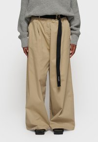 Pantalons larges beige en tissu lisse, dotés d'une ceinture noire avec une boucle en argent et de poches latérales visibles.
