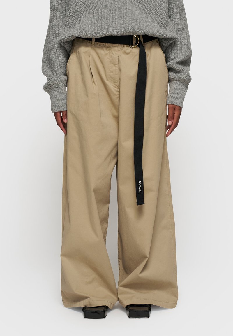 Pantalons larges beige en tissu lisse, dotés d'une ceinture noire avec une boucle en argent et de poches latérales visibles.