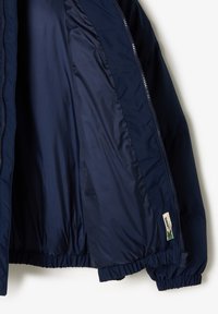 Veste isolée bleu marine avec une doublure intérieure brillante, des poignets élastiques et une étiquette logo sur l'ourlet. Fermeture zippée sur toute la longueur et design bouffant.