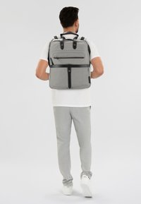 Zaino grigio in tessuto con dettagli in pelle nera, dotato di spallacci regolabili e una tasca anteriore con zip. Design minimalista.