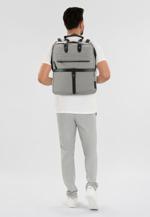 Zaino grigio in tessuto con dettagli in pelle nera, dotato di spallacci regolabili e una tasca anteriore con zip. Design minimalista.