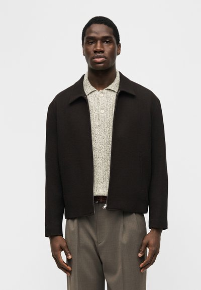 Νέος άνδρας που φοράει μαύρο ζακέτα με φερμουάρ, ελαφρώς υφασμένο πόλο πουκάμισο και γκρι παντελόνι, στέκεται μπροστά σε ένα απλό φωτεινό φόντο.