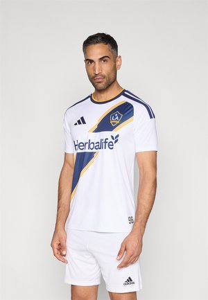 Weißes Fußballtrikot mit blauem und gelbem diagonalen Streifen, "Herbalife"-Logo und LA Galaxy-Wappen. Mit kurzen Ärmeln und rundem Ausschnitt.