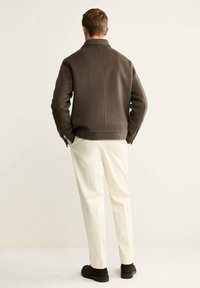 Giacca in lana marrone con colletto alto, polsini a coste e design pulito, abbinata a pantaloni crema e scarpe scure.