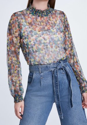 Frau trägt eine transparente, florale Langarmbluse mit Rüschenkragen und -manschetten, kombiniert mit hoch taillierten blauen Denim-Jeans mit gebundenem Gürtel.