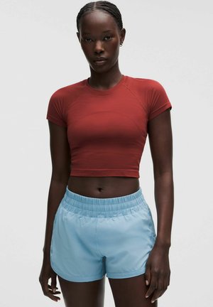 Vrouw draagt een nauwsluitende, kortermige rusty cropped top en lichtblauwe elastische sportshorts, staande tegen een effen achtergrond.