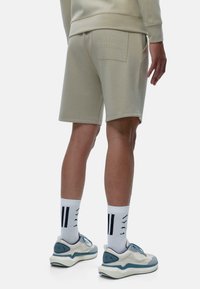 Shorts en coton beige avec une poche arrière comportant un texte en relief. Porté avec des chaussettes blanches et des baskets à accents bleus. Texture lisse, coupe décontractée.