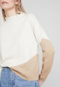 Jersey de punto con cuello alto, en bloque de colores blanco y beige, con textura suave y un corte relajado. Mangas largas con hombros caídos.