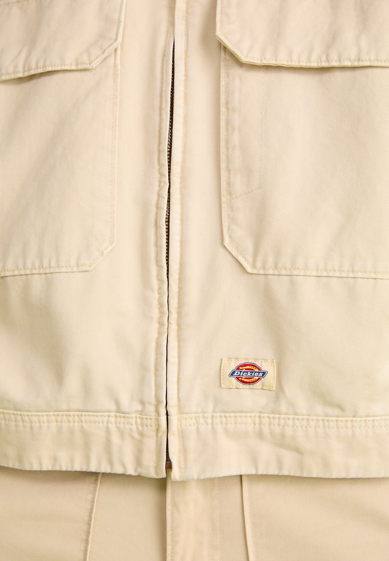 Veste de travail beige en tissu résistant, dotée d'un zip sur le devant, de deux poches poitrines et d'une petite étiquette colorée au bas de l'ourlet.
