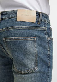 Džínové kalhoty v středně modré barvě s vybledlým vzhledem, s oranžovým prošíváním, koženou štítkem na pasu a dvěma zadními kapsami.