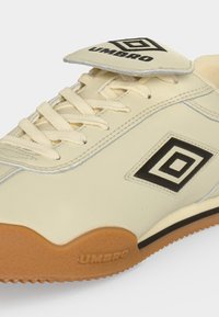 Μπεζ sneaker Umbro με κάλυμμα κορδονιών, μαύρο διπλό διαμαντένιο λογότυπο και καφέ λαστιχένια σόλα, που δείχνει πλαϊνή και πάνω όψη.