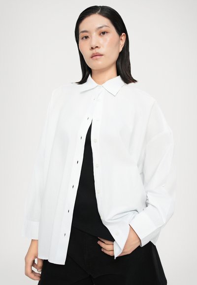 Λευκό πουκάμισο σε υπερμεγέθη γραμμή με κλασικό γιακά, μαύρα κουμπιά και μακριά μανίκια. Φορεμένο πάνω από μαύρο τοπ, με μινιμαλιστικό σχεδιασμό.