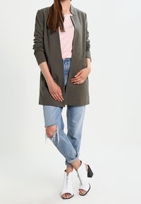 Manteau long vert olive, haut rose clair, jeans bleu clair déchirés, et bottes blanches à zip et talon bloc.