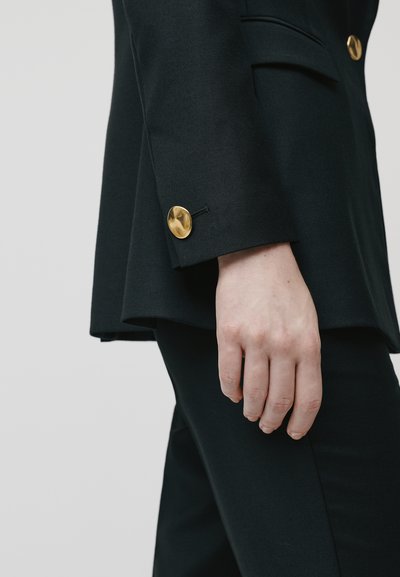 Musta bleiseri, jossa on väljempi malli ja kultanappi hihassa. Kangas näyttää sileältä ja siinä on jämäkkää muotoilua hihojen ympärillä.