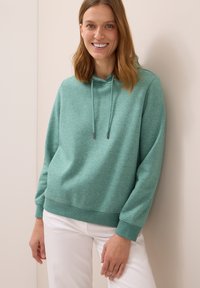 Tealfärgad hoodie av mjukt, texturerat tyg, med dragsko i huvan och ribbade ärmslut, kombineras med vita byxor för kontrast.