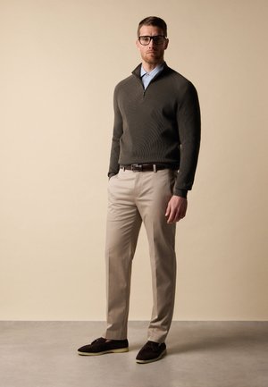 Uomo con occhiali neri, maglione scuro con zip al collo, camicia azzurro chiaro, pantaloni beige e mocassini scuri, in piedi su uno sfondo beige.