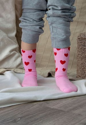 Nogi dziecka w szarych spodniach i różowych skarpetkach z czerwonymi serduszkami stojące na białym kocu na drewnianej podłodze obok plecionej koszyczka.