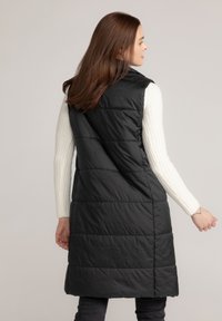 Zwarte gewatteerde vest met een hoge kraag, voorzien van verticale stiksels en een losse pasvorm, gedragen over een witte geribbelde trui.