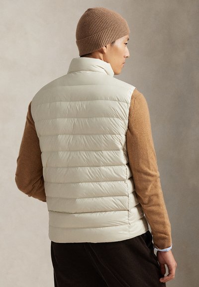 Ljusbeige vadderad väst med horisontell sömnad, hög krage och slät textur, bärs över en brun tröja. Ribbad brun mössa på huvudet.
