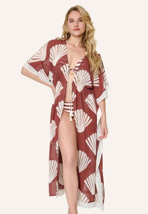 Blonde vrouw staat met hand op heup, draagt een roestkleurige open cover-up met witte schelpenprint en een beige bikini eronder.