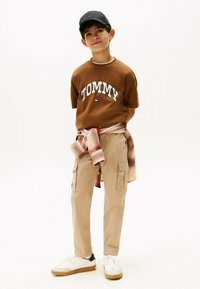 Bruine katoenen T-shirt met "TOMMY" logo, beige cargo pants, grijze en oranje flanellen shirt om de taille gebonden, zwarte pet, witte sneakers met beige zolen.