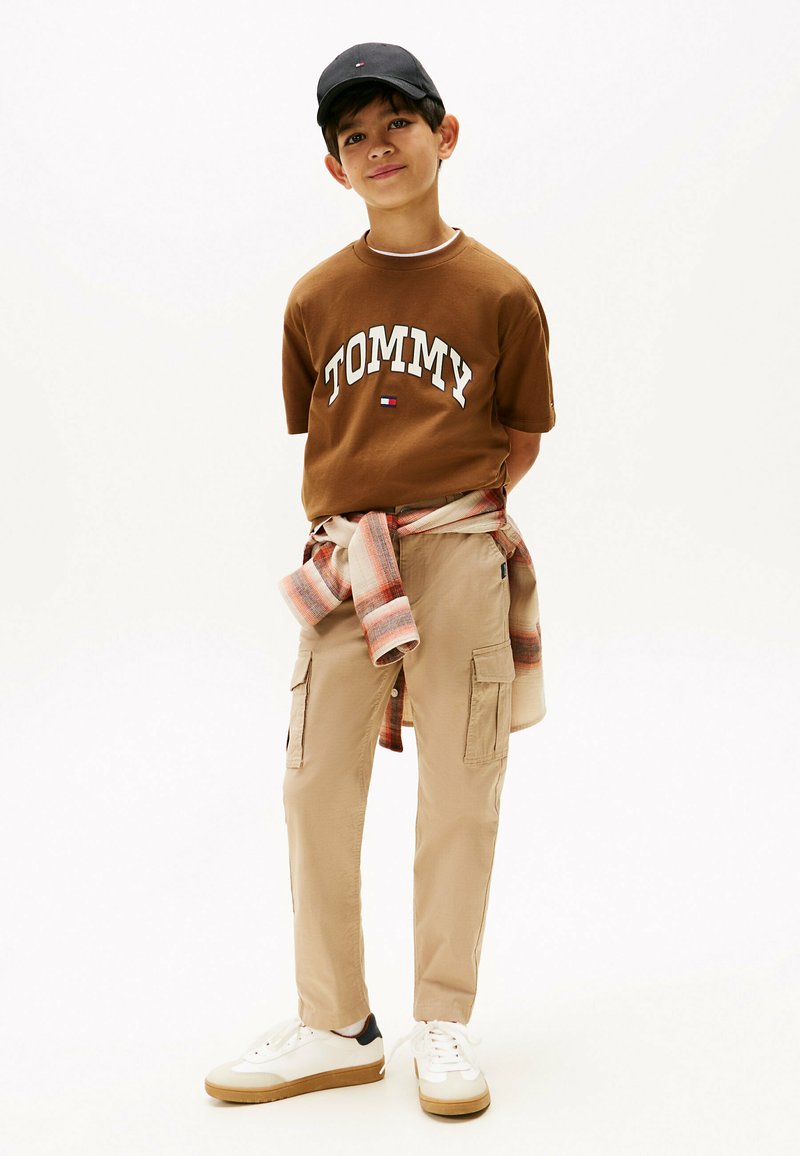 Bruine katoenen T-shirt met "TOMMY" logo, beige cargo pants, grijze en oranje flanellen shirt om de taille gebonden, zwarte pet, witte sneakers met beige zolen.