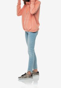 Felpa rosa chiaro con tasca frontale e lacci, abbinata a jeans skinny blu chiaro e sneaker nere con stampa a serpente.