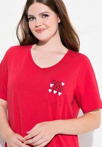 T-shirt en coton rouge à col en V avec des manches courtes, comportant une poche affichant un motif de jeu de morpion avec des "X" et des cœurs en noir et blanc.