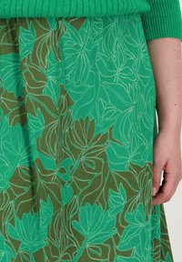 Jupe florale verte avec un motif de feuilles souligné de blanc sur un fond texturé marron, dotée d'un design plissé et d'une taille élastique.