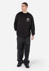 Sweatshirt noir à col rond associé à un pantalon en denim foncé ample, avec des coutures contrastées et des designs de poches uniques. Des baskets noires complètent le look.